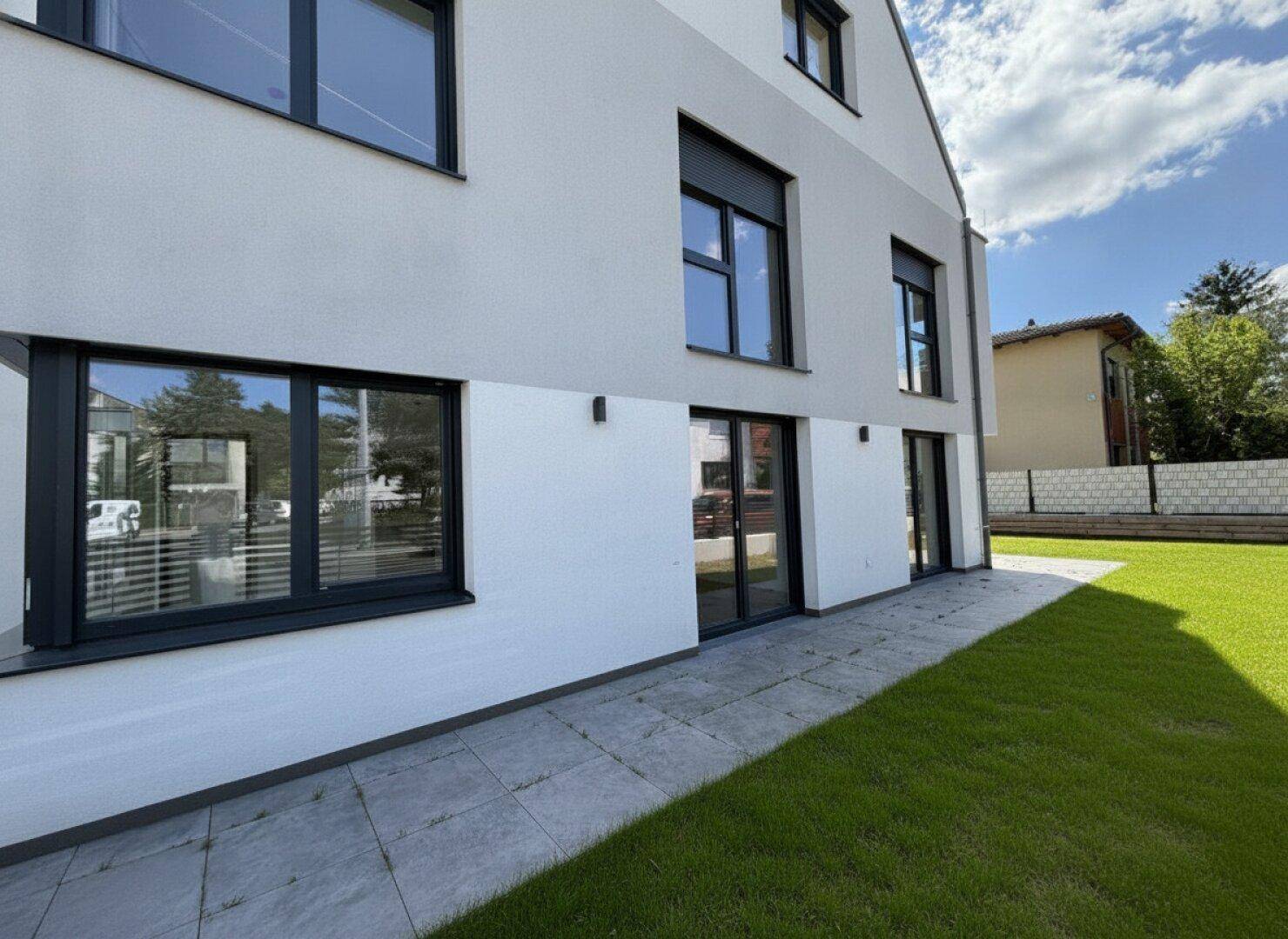 Moderne Gebäudefassade mit großen Fenstern, Rollläden und einer gepflegten Rasenfläche.