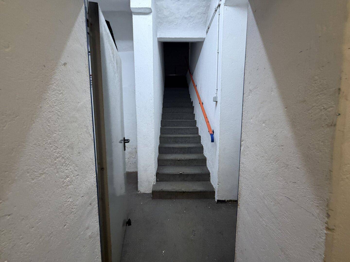Schmaler Treppenabgang mit Betonstufen und einem orangefarbenen Handlauf, der in einen dunklen Bereich führt.