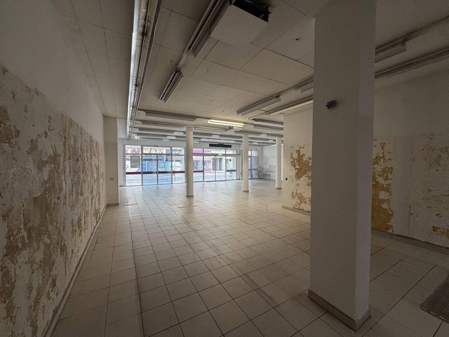 Weitwinkelaufnahme des Geschäftsraums mit Fliesenboden, Säulen und großen Schaufenstern zur Straße.
