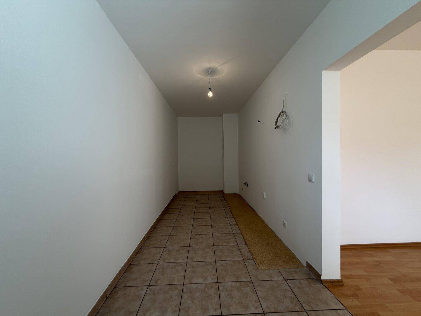 Langer, gefliester Raum mit weißen Wänden und vorbereiteten Anschlüssen, ideal für eine Küche.