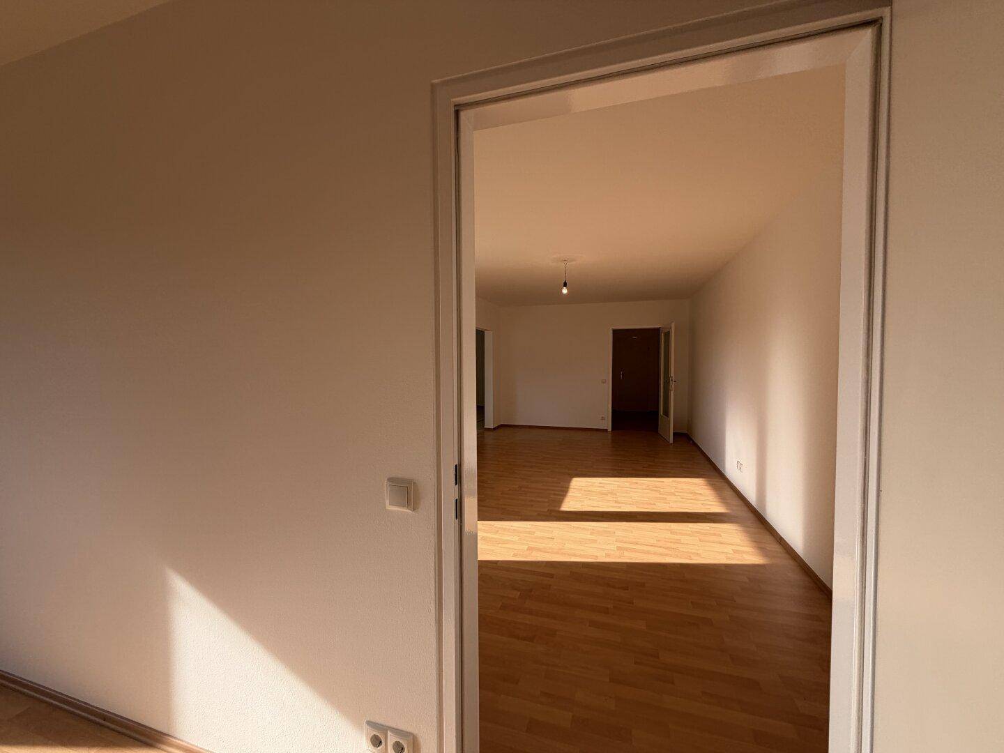 Blick durch eine offene Tür in einen hellen Raum mit Laminatboden, der von Sonnenlicht durchflutet wird.