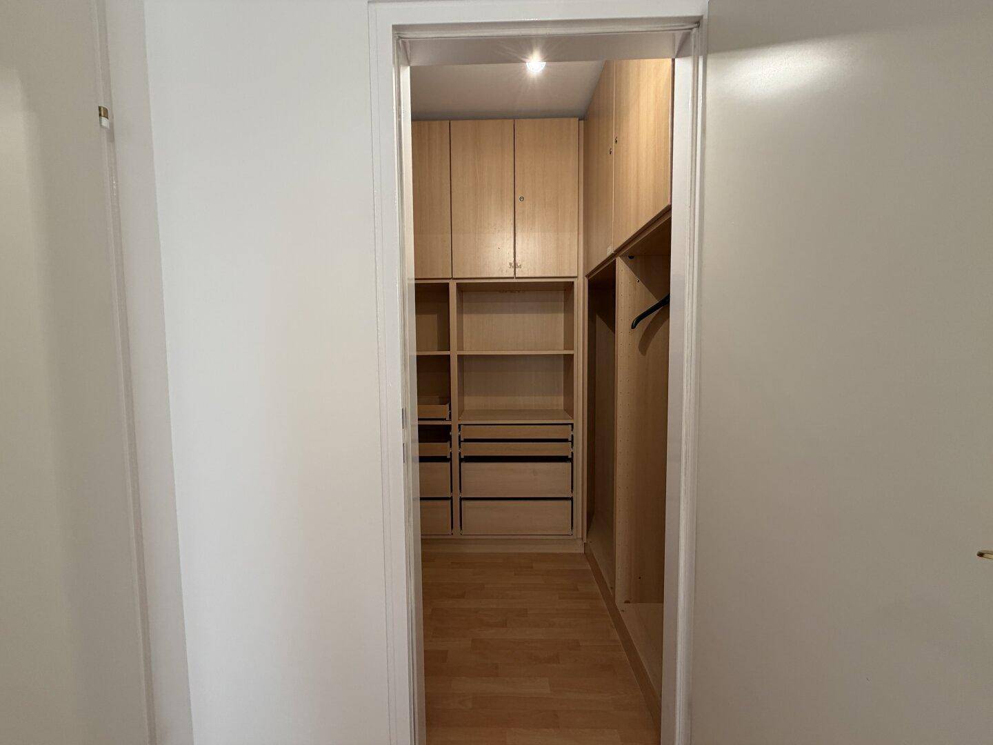 Blick durch eine Tür in einen begehbaren Kleiderschrank mit Holzregalen und Stauraum.