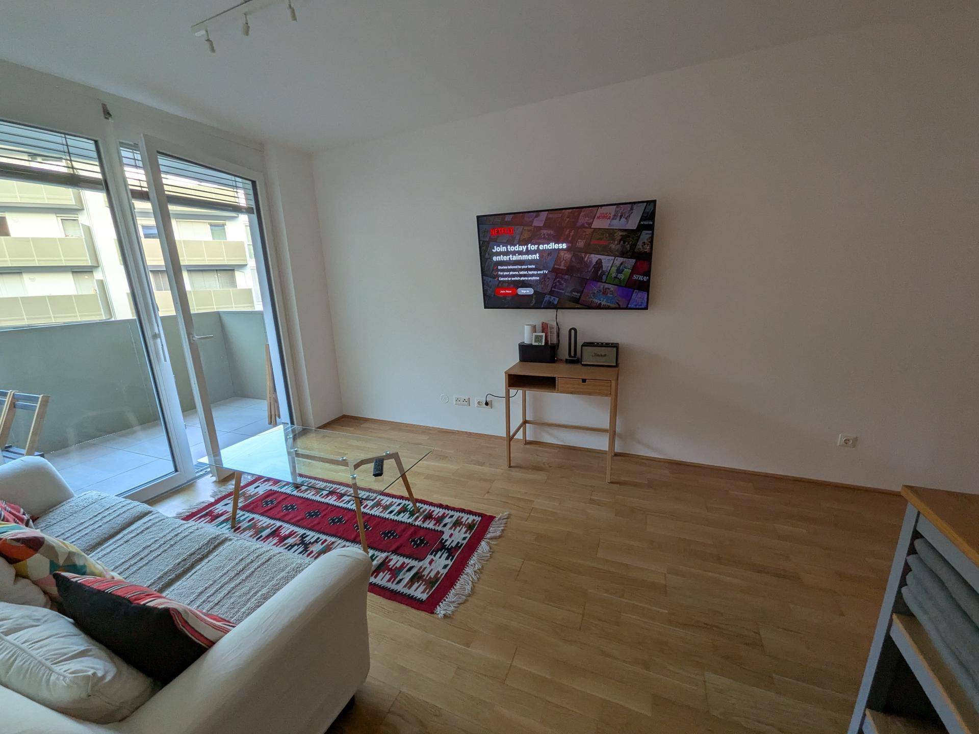 Gemütliches Wohnzimmer mit Sofa, Fernseher und großen Fenstern zum Balkon.