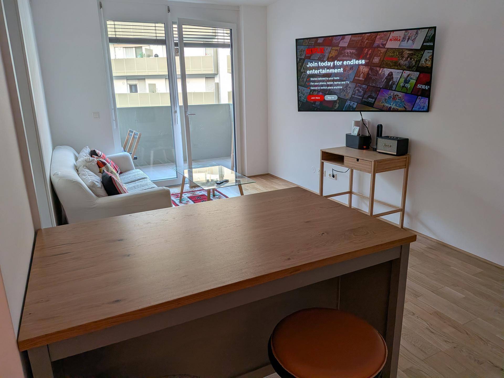Heller Wohnbereich mit Esstisch, Sofa, Fernseher und Zugang zum Balkon.