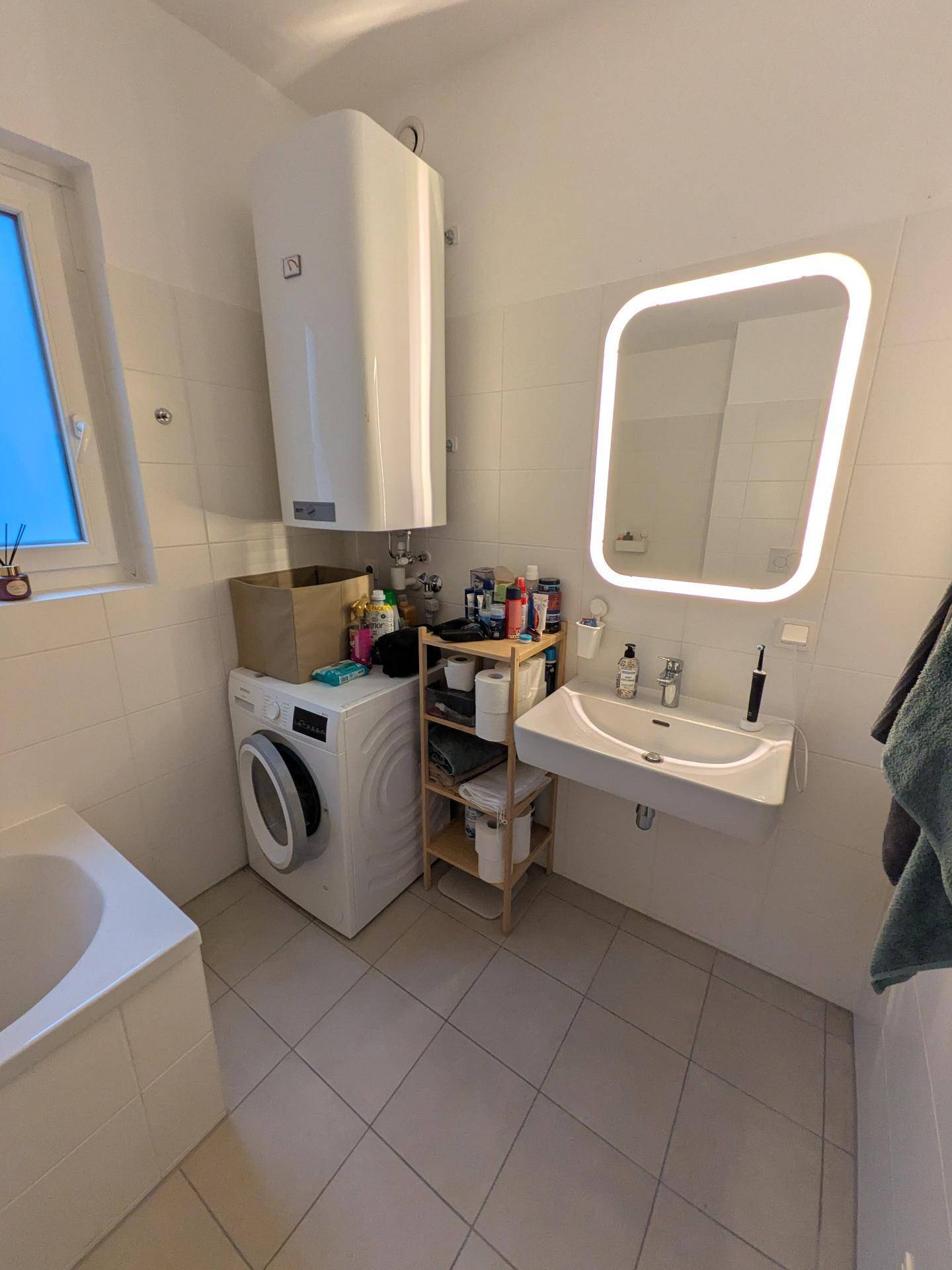 Helles Badezimmer mit Waschbecken, beleuchtetem Spiegel, Waschmaschine und Fenster.