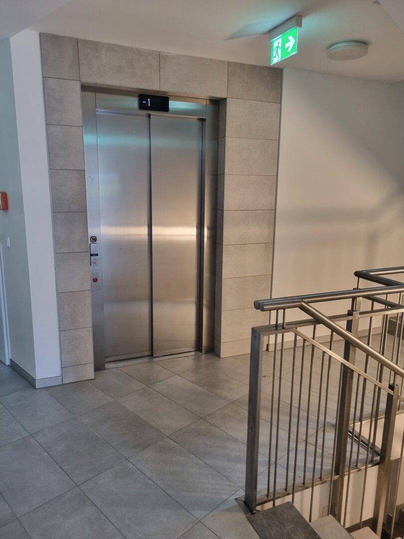 Moderner Aufzug in einem hellen Flur mit Fliesenboden und Treppenhauszugang.