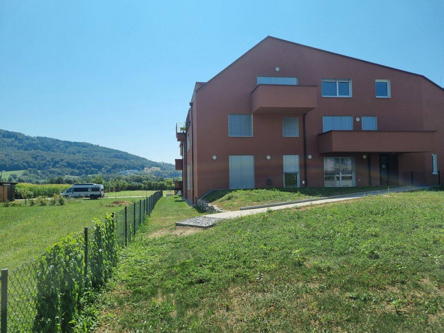 Seitenansicht des Wohngebäudes mit roter Fassade, umgeben von Feldern und Hügeln.