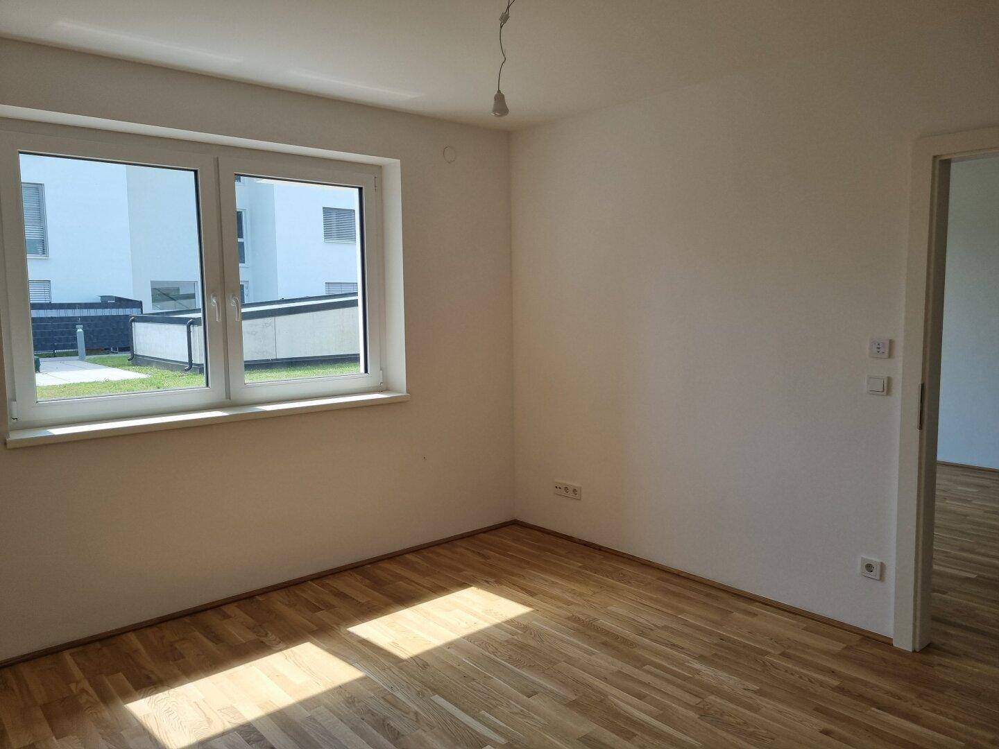 Unmöbliertes Zimmer mit Holzboden und Fenster, das viel Tageslicht hereinlässt.