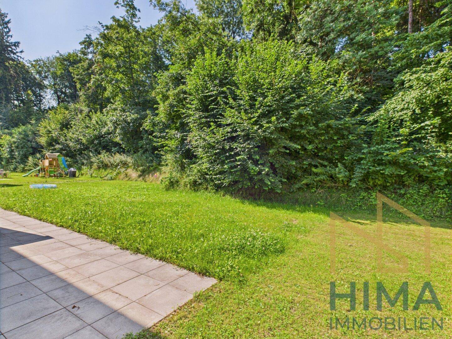 Großer Gartenbereich mit Rasenfläche, gepflasterter Terrasse und Spielplatz im Hintergrund.