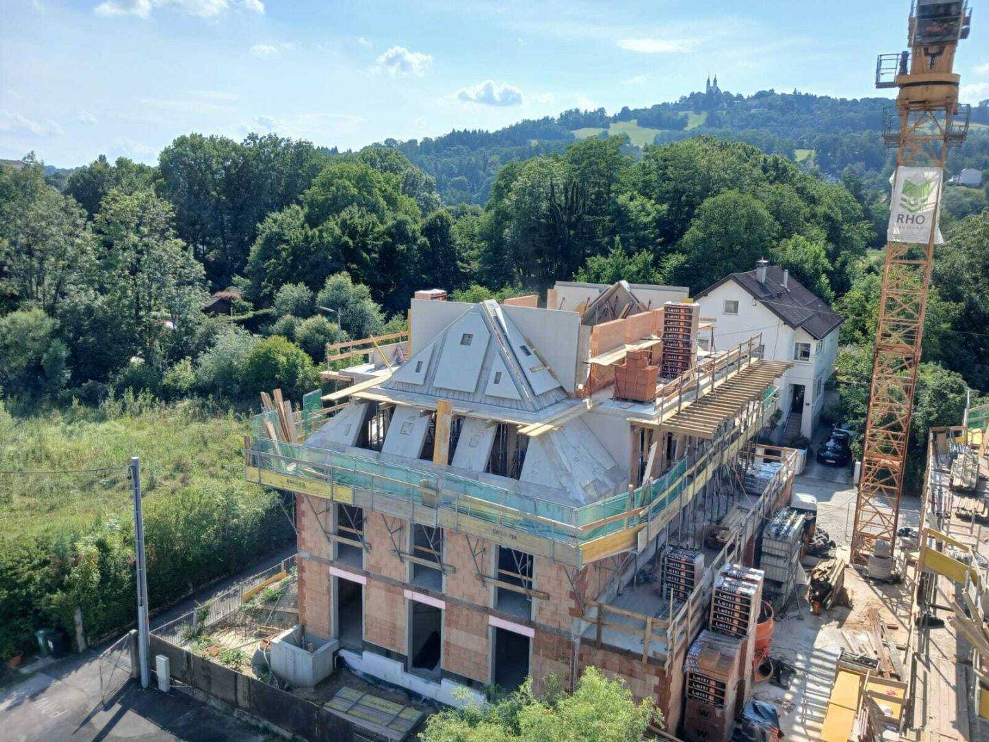 Seitenansicht eines im Bau befindlichen Gebäudes mit sichtbaren Dachstrukturen und grüner Umgebung.