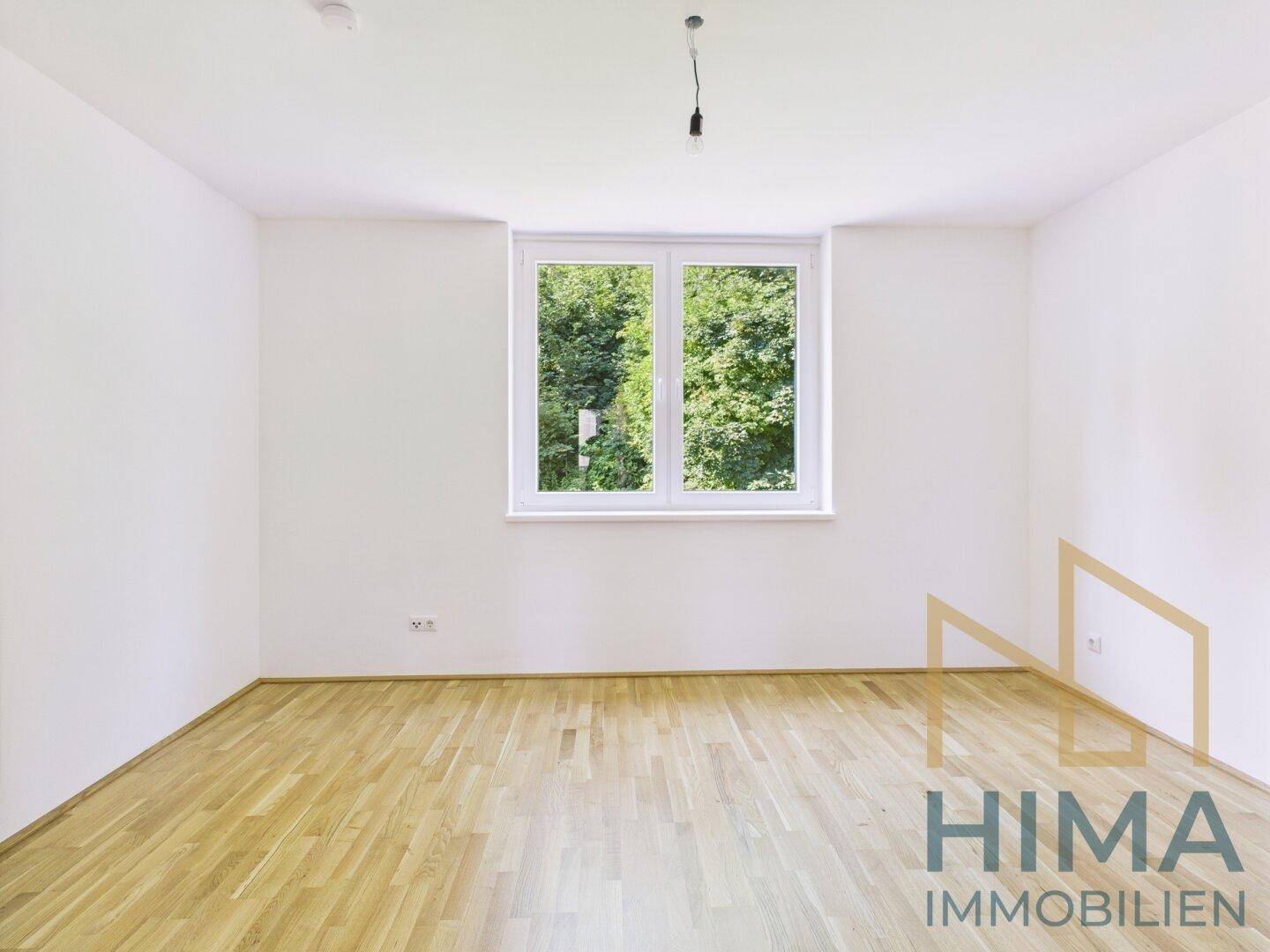 Leeres Zimmer mit Parkettboden und Fenster mit Blick ins Grüne.