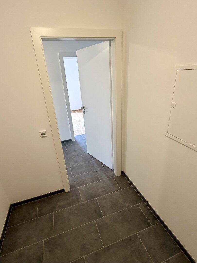 Heller Flur mit dunklen Fliesen und einer offenen Tür, die den Blick in einen weiteren Raum freigibt.