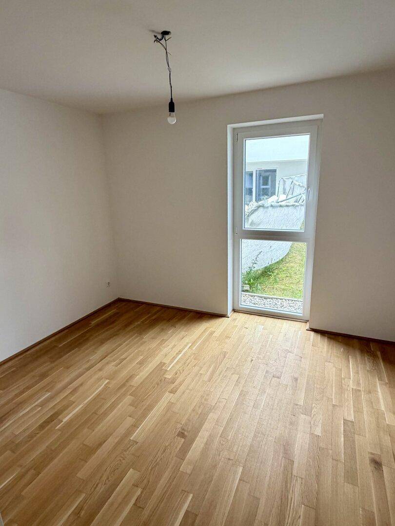Helles Schlafzimmer mit Parkettboden und einem Fenster, das den Blick auf eine Grünfläche freigibt.