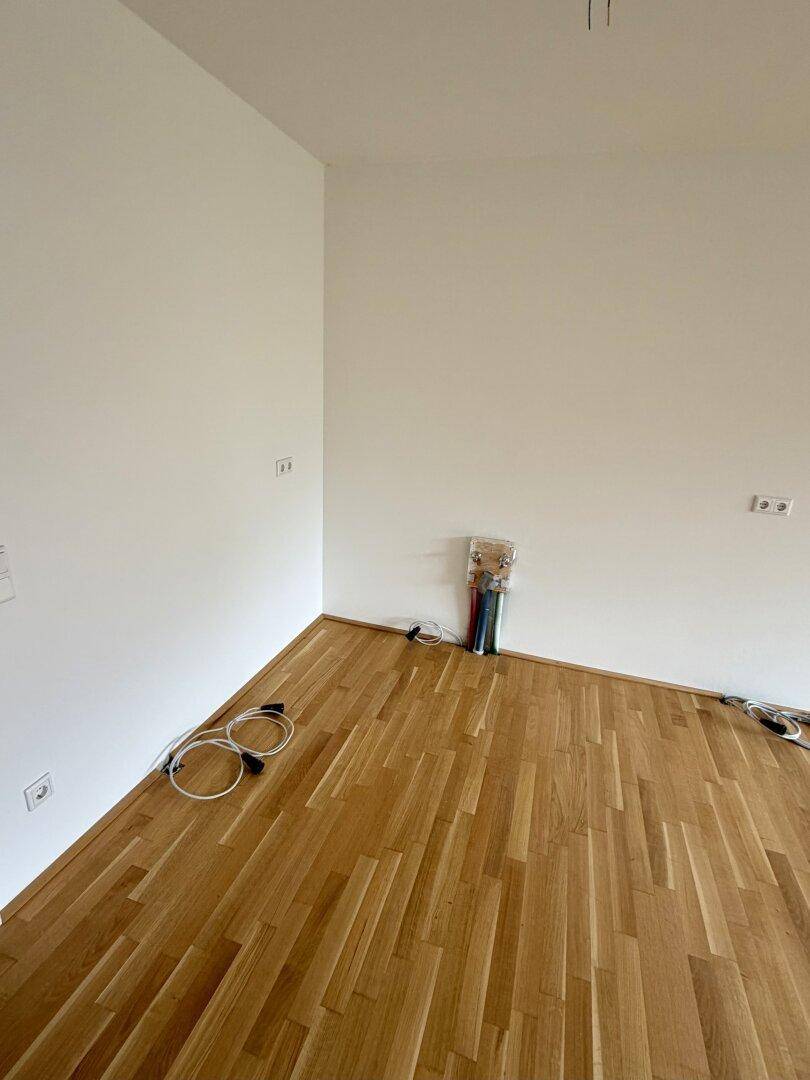 Ecke eines leeren Raumes mit Parkettboden und sichtbaren Elektroinstallationen, bereit für die Einrichtung.