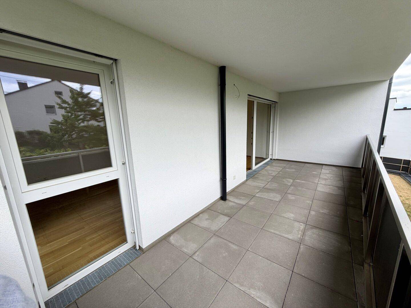 Langer, gefliester Balkon mit weißer Fassade und Blick auf die umliegenden Gebäude und Bäume.