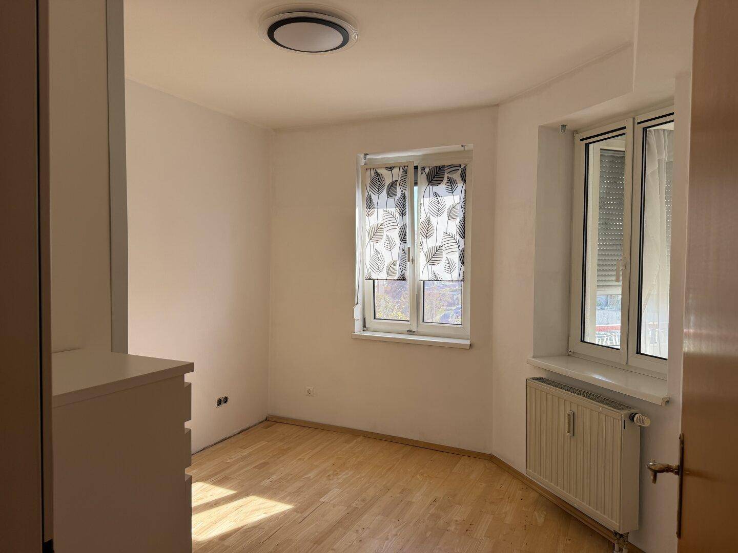 Helles Schlafzimmer mit Laminatboden, weißen Wänden und Fenster mit gemustertem Vorhang.