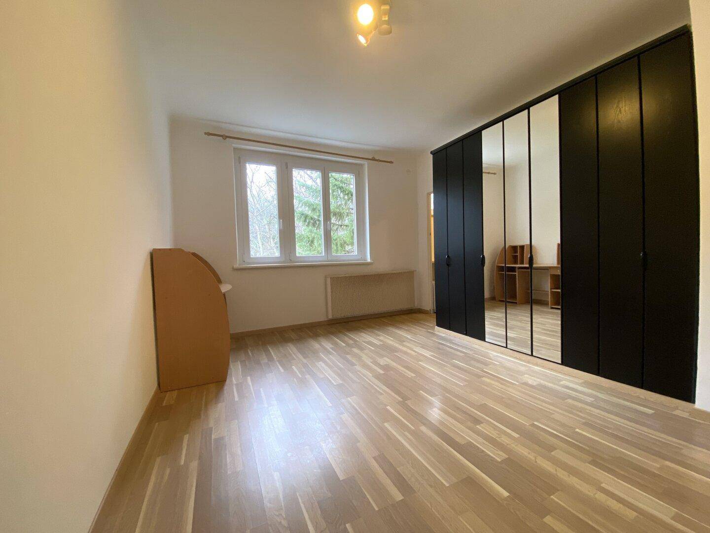 Geräumiges Zimmer mit Holzfußboden, Fenster und einem großen Kleiderschrank mit Spiegeltüren.