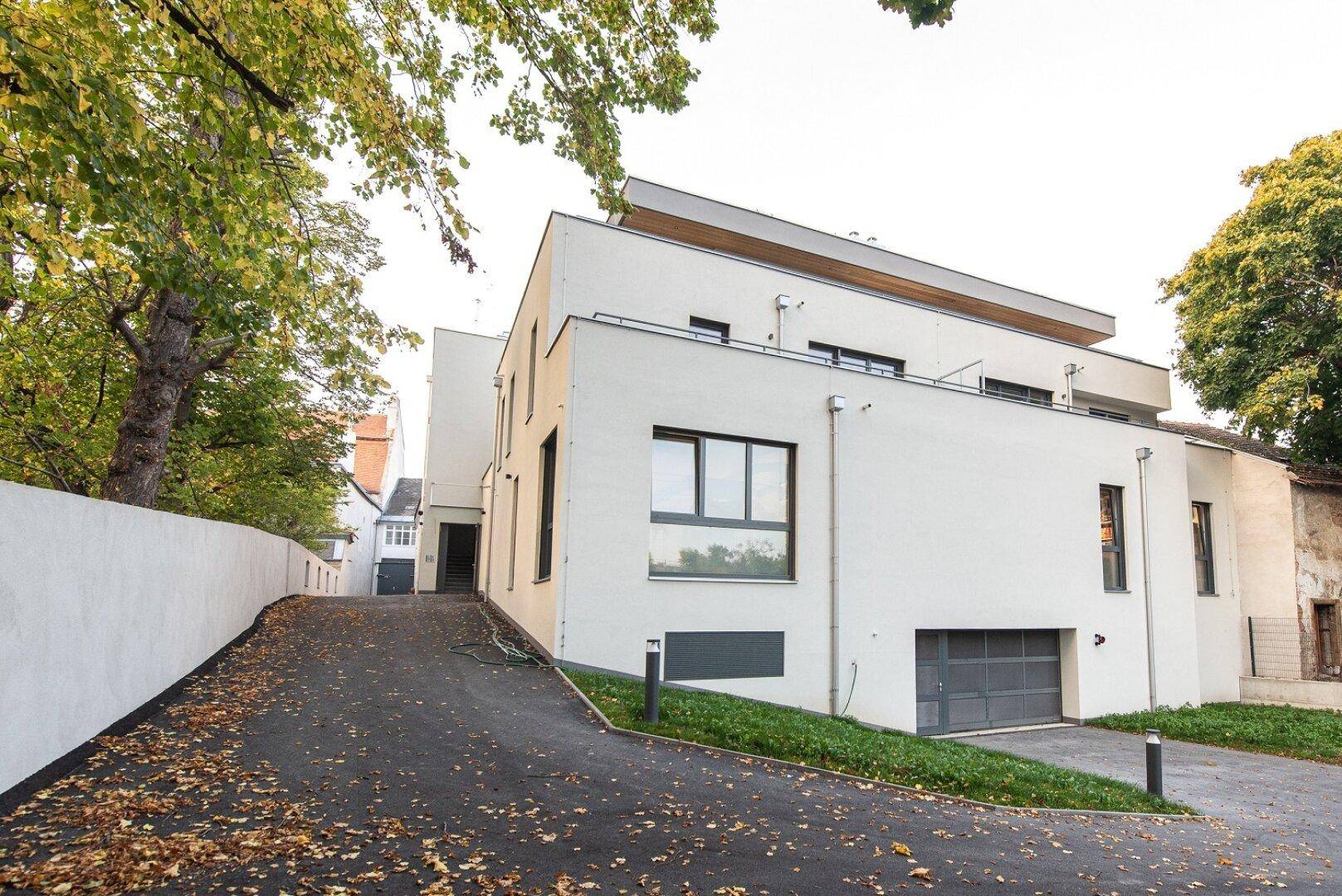 Modernes Gebäude mit Zufahrt zur Garage, umgeben von Bäumen und Grünflächen.