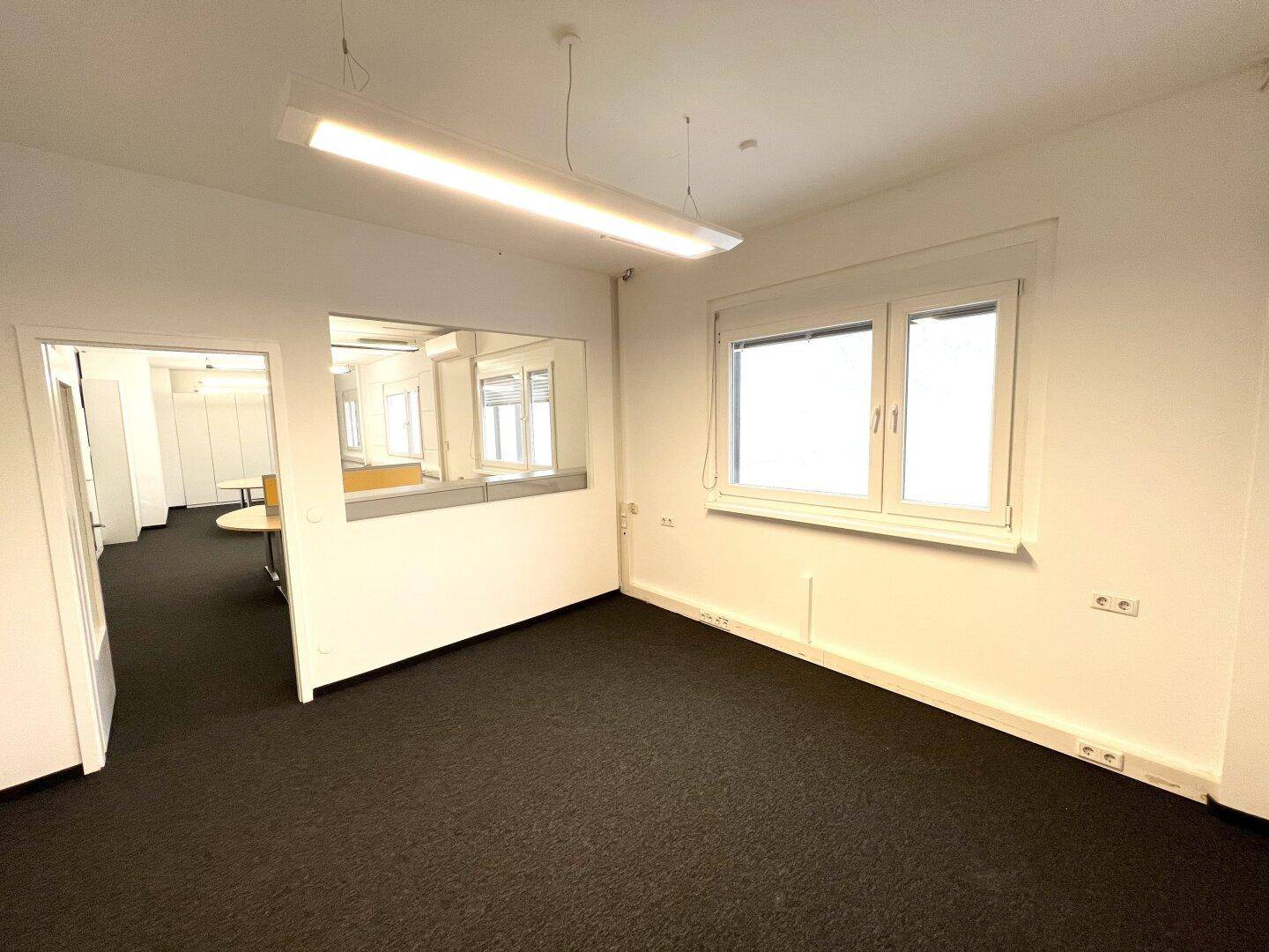 Heller Büroraum mit dunklem Teppichboden, einem Fenster und einer Trennwand mit integriertem Glasausschnitt.