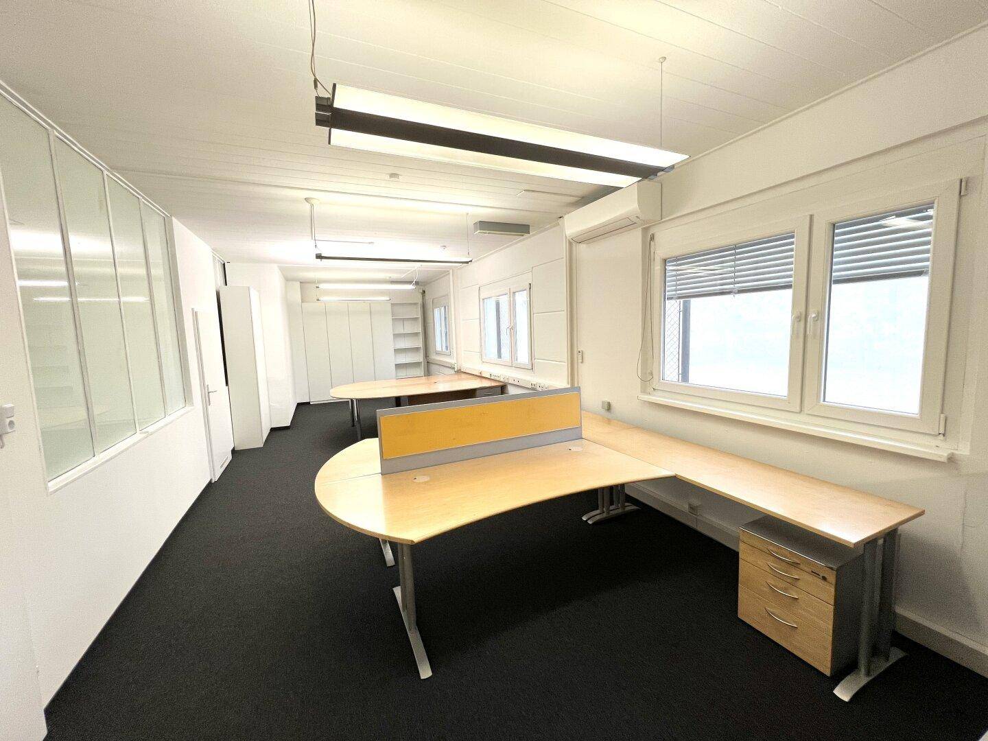 Geräumiger Büroraum mit mehreren Arbeitsplätzen, Teppichboden und großen Fenstern mit Jalousien.