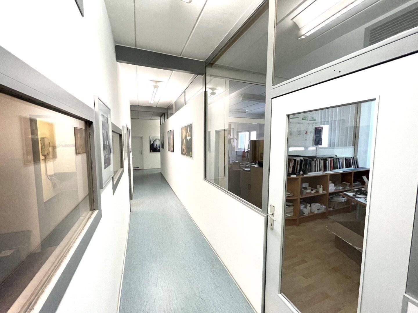 Heller Flur mit Teppichboden, Glaswänden zu Büros und gerahmten Bildern an den Wänden.