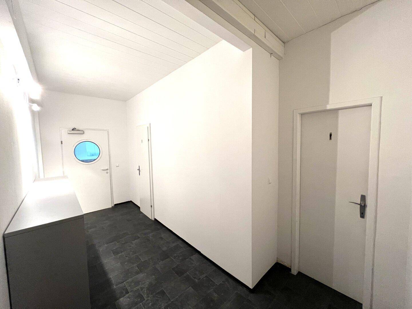 Schlichter Flur mit dunklem Fliesenboden und mehreren weißen Türen, darunter eine mit einem runden Fenster.