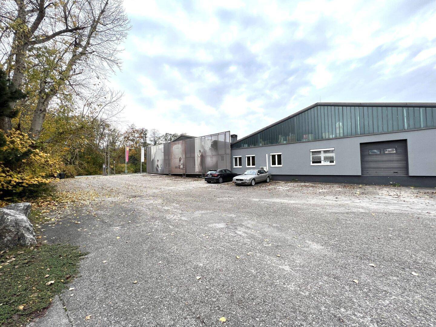 Außenansicht eines Gewerbegebäudes mit großem Schotterparkplatz und umliegenden Bäumen.