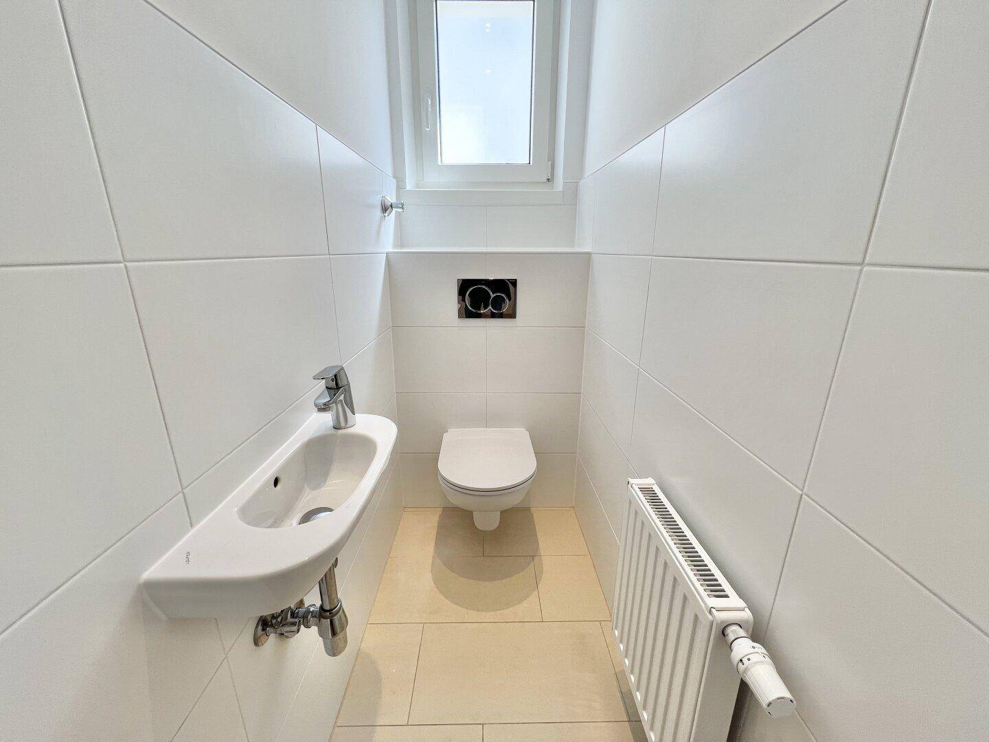 Separates WC mit wandhängender Toilette, kleinem Waschbecken und Fenster für natürliche Belüftung.