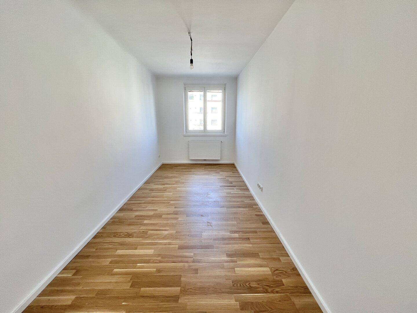 Langes, schmales Zimmer mit Parkettboden und einem Fenster, geeignet als Kinder- oder Gästezimmer.