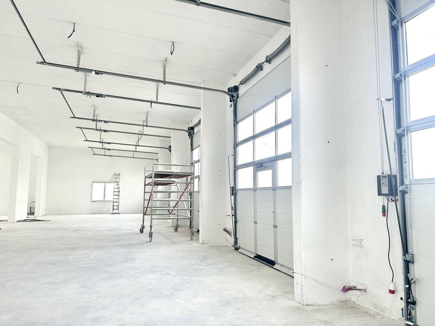 Blick in eine große, helle Industriehalle mit hohen Decken, einem Rolltor und einem Gerüst im Vordergrund.