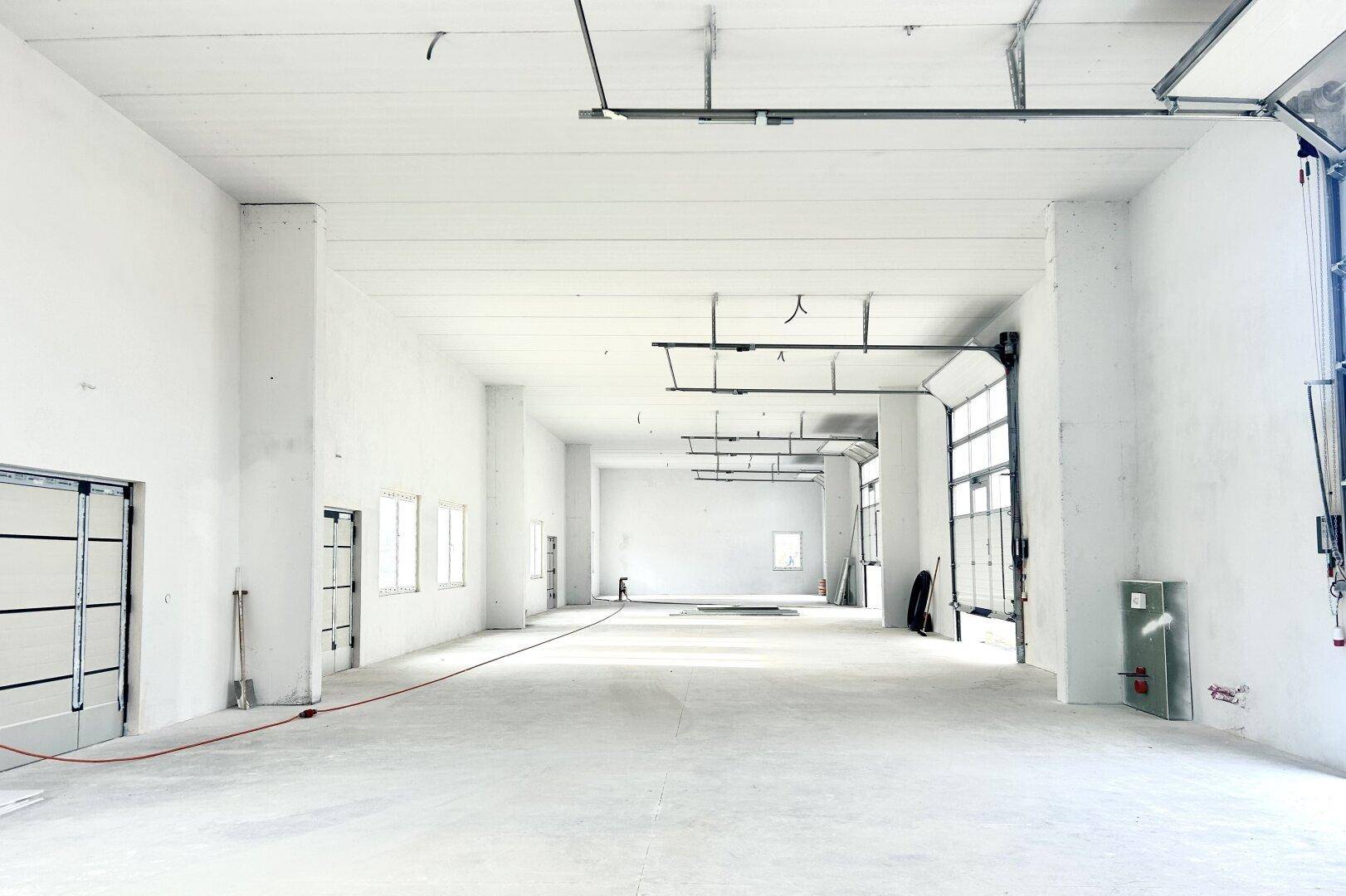 Geräumige, leere Halle mit hohen Decken, Betonboden und großen Rolltoren, die viel Tageslicht hereinlassen.