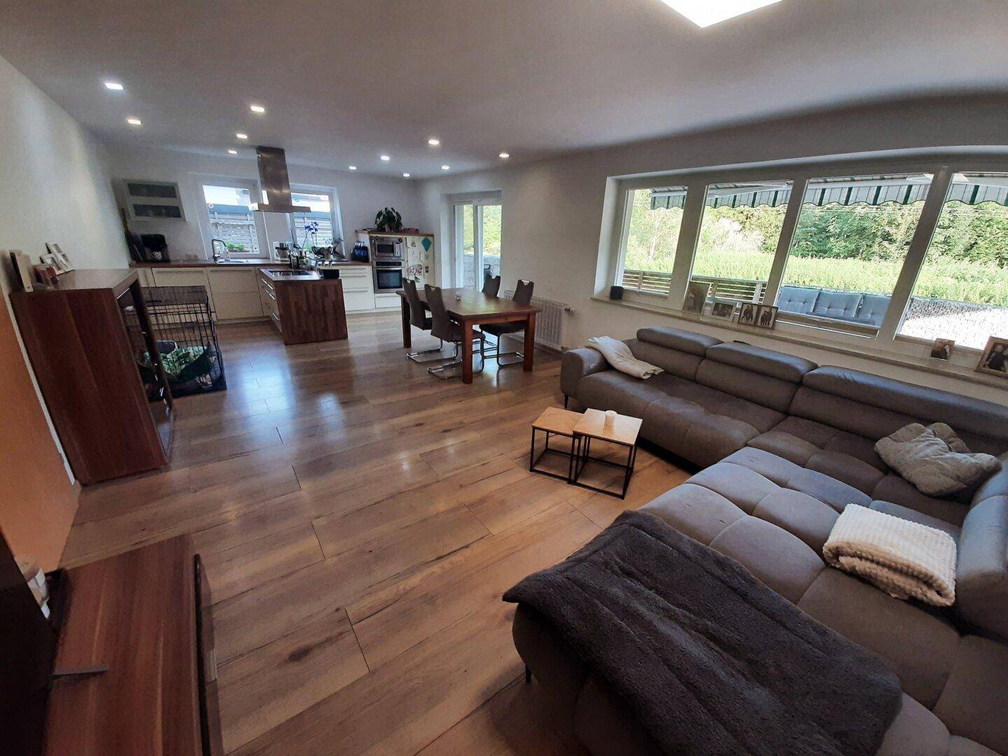 Offener Wohn-Essbereich mit Holzboden, moderner Küche und großen Fenstern mit Grünblick.