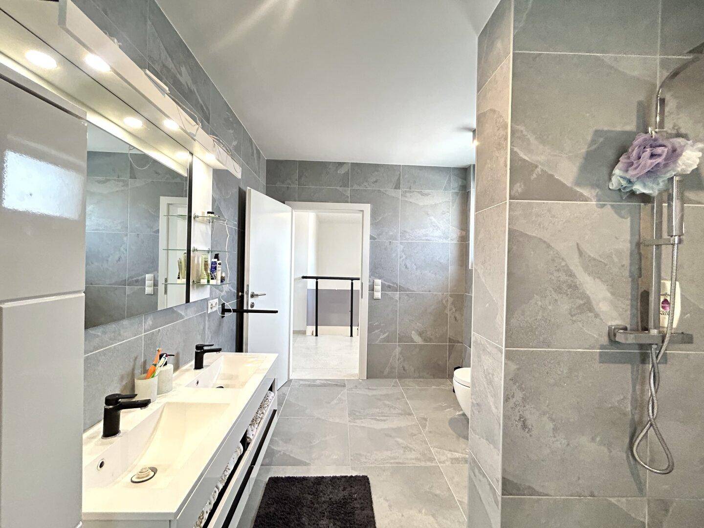 Helles, modernes Badezimmer mit Doppelwaschbecken, großer Dusche und grauen Fliesen.