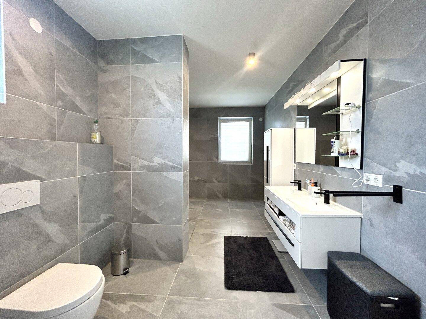 Modernes Badezimmer mit großen grauen Fliesen, Doppelwaschbecken und wandhängendem WC.
