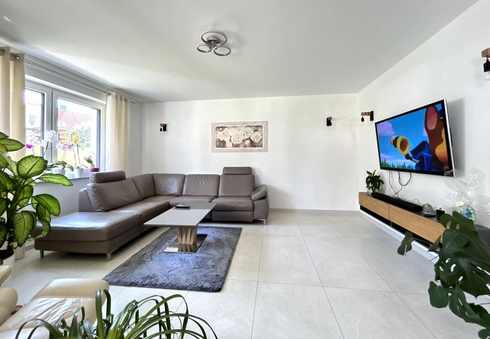 Geräumiges Wohnzimmer mit beigem Ecksofa, Fliesenboden und einem großen Fenster.