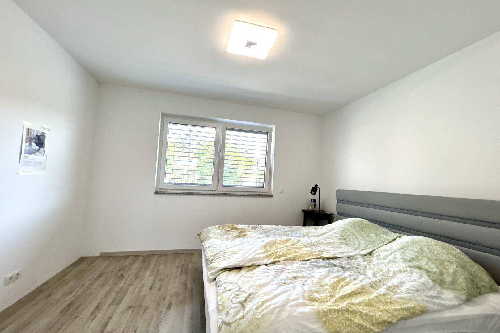 Schlafzimmer mit Laminatboden, großem Fenster und einem Doppelbett mit Kopfteil.