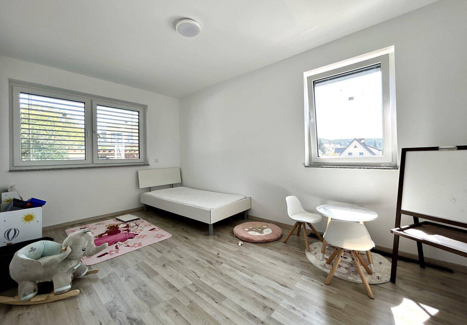 Kinderzimmer mit Laminatboden, zwei Fenstern und einem Einzelbett sowie Spielbereich.