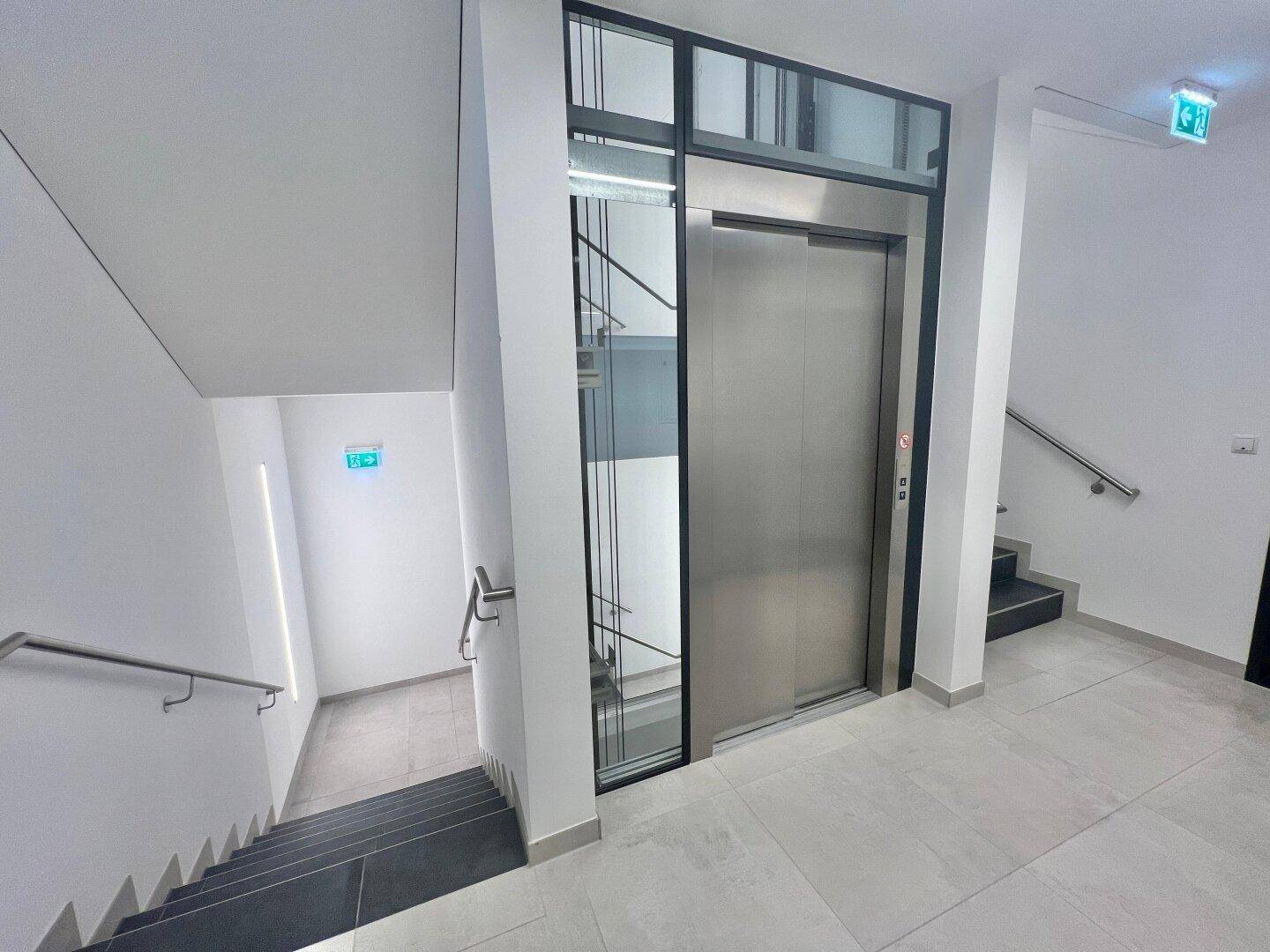 Moderner Aufzug mit Edelstahltüren und Glasfront im Treppenhaus des Gebäudes.