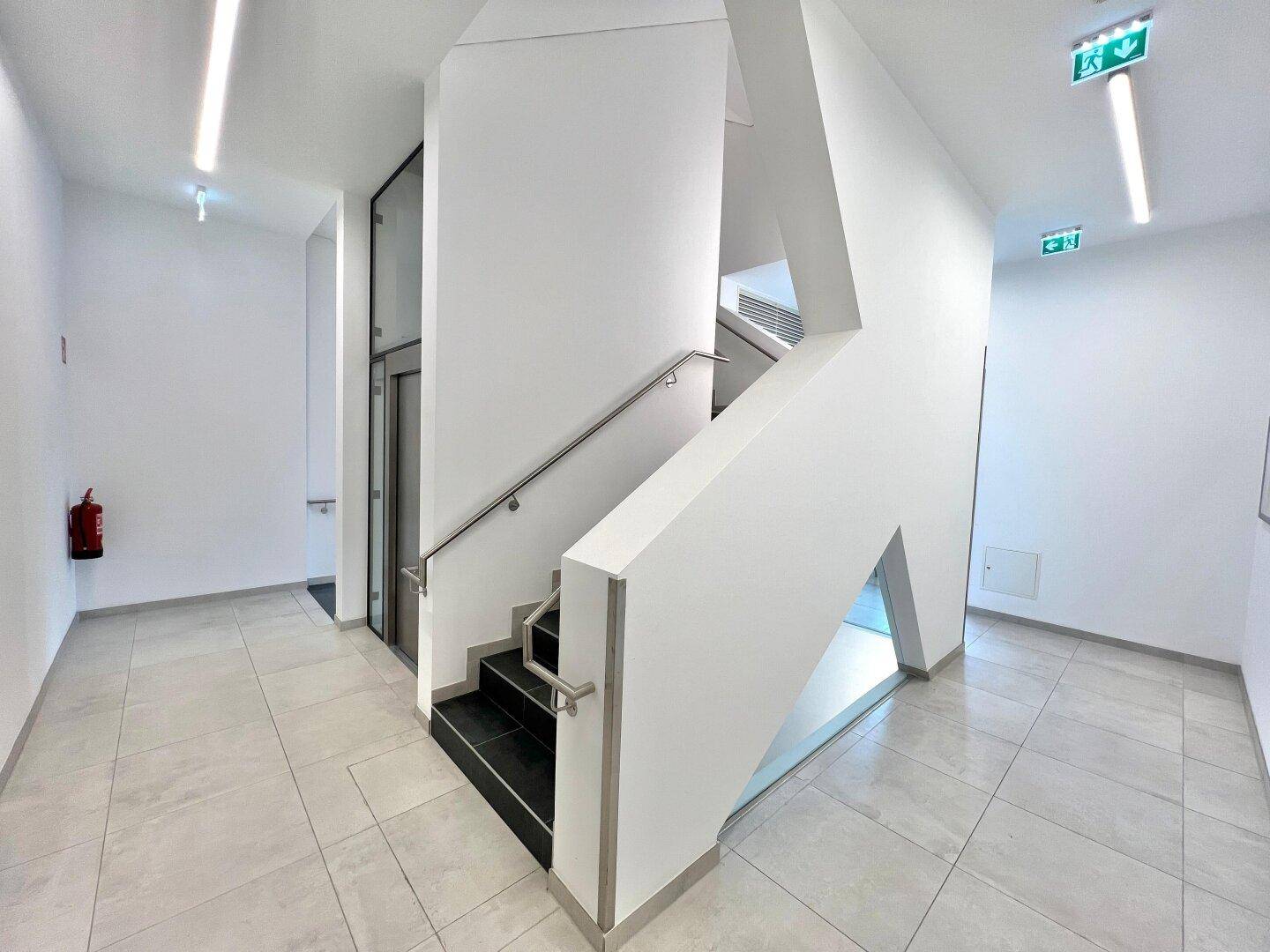 Helles Treppenhaus mit modernem Design, Edelstahlgeländer und Zugang zum Aufzug.