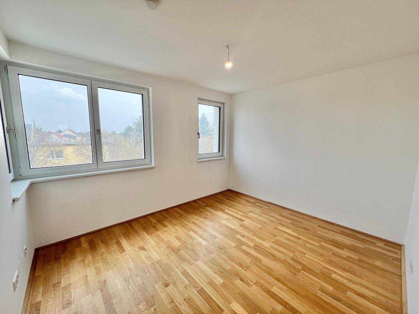 Helles, leeres Zimmer mit Parkettboden und großen Fenstern, die viel Tageslicht hereinlassen.