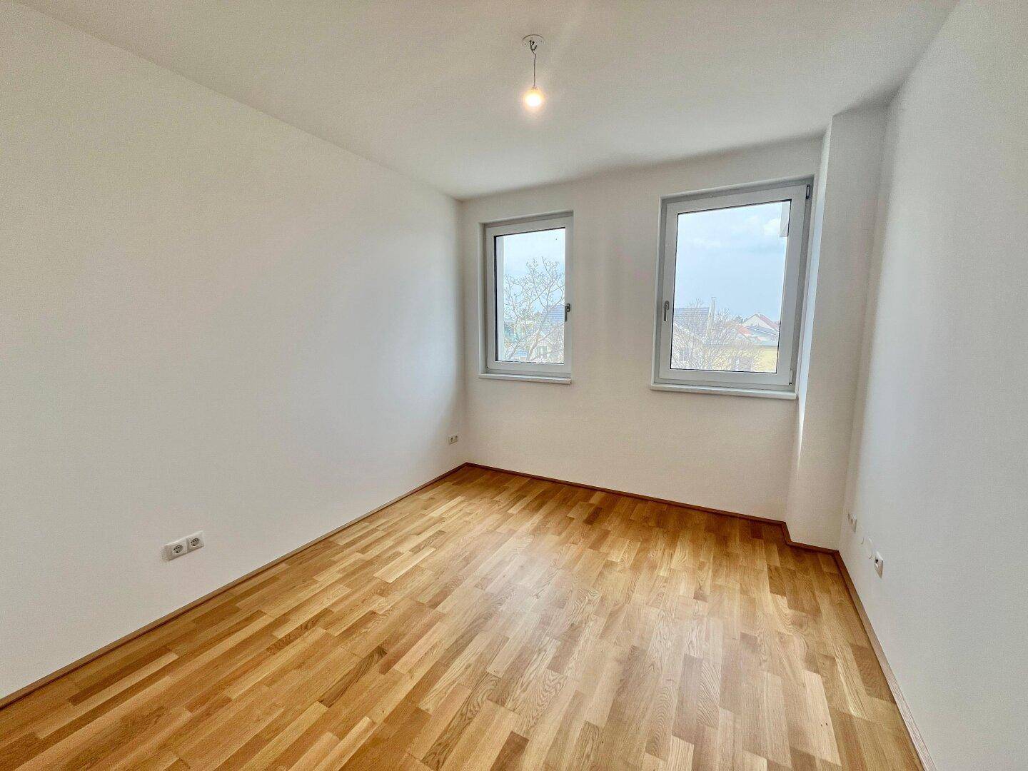 Unmöbliertes Zimmer mit Parkettboden und zwei großen Fenstern, die den Raum erhellen.