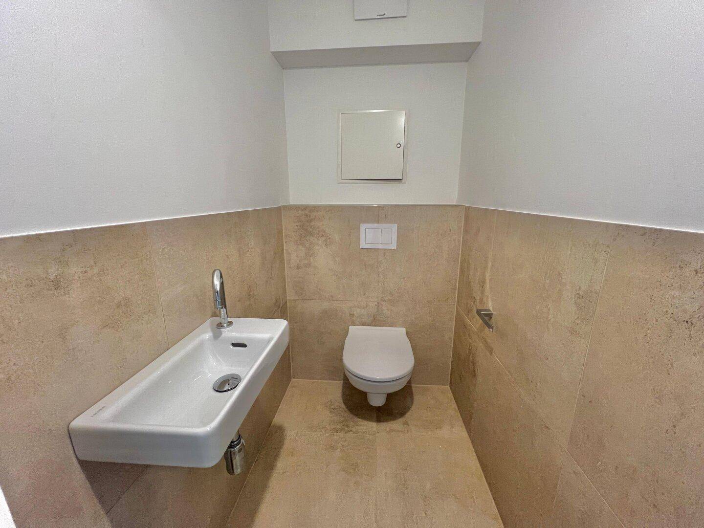 Gäste-WC mit wandhängendem WC und kleinem Waschbecken, halbhoch gefliest in hellen Beigetönen.