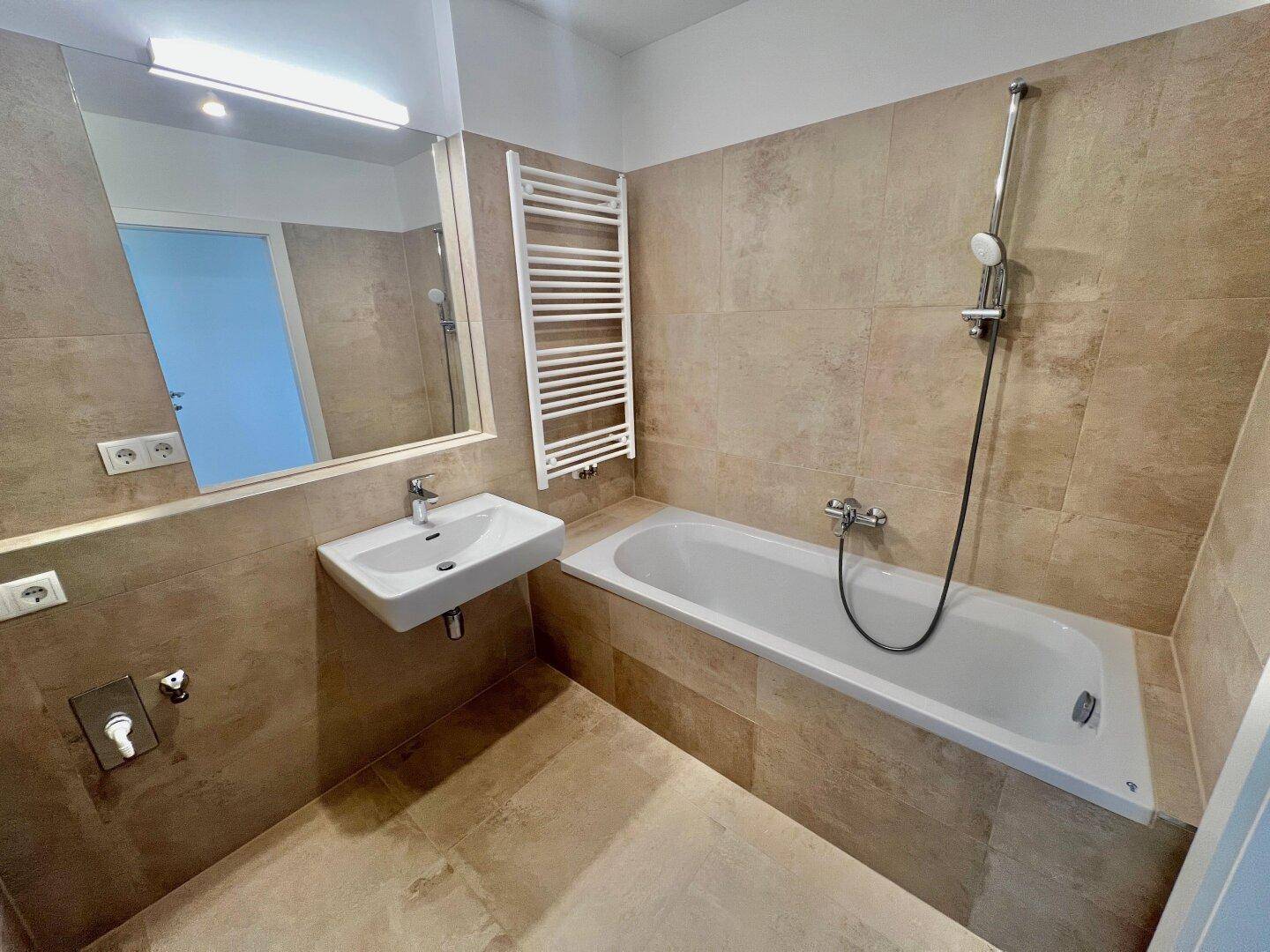 Modernes Badezimmer mit Badewanne, Handbrause und beheiztem Handtuchhalter, gefliest in hellen Beigetönen.
