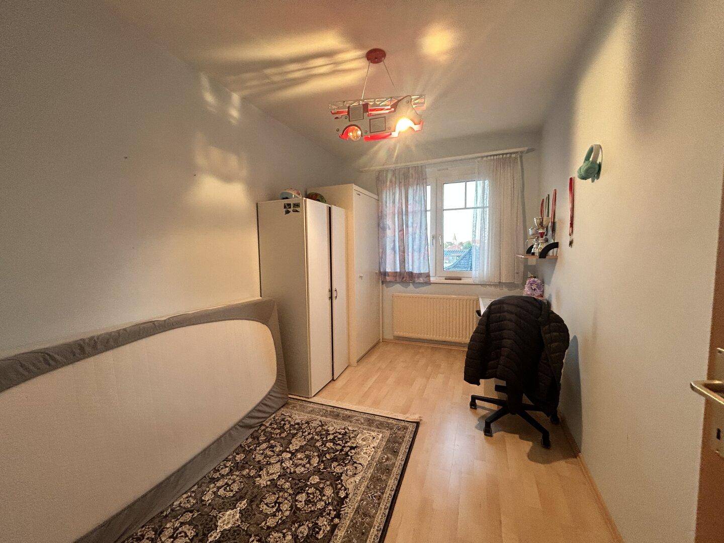 Helles Zimmer mit Schreibtisch, Kleiderschrank und Fenster mit Blick nach draußen.