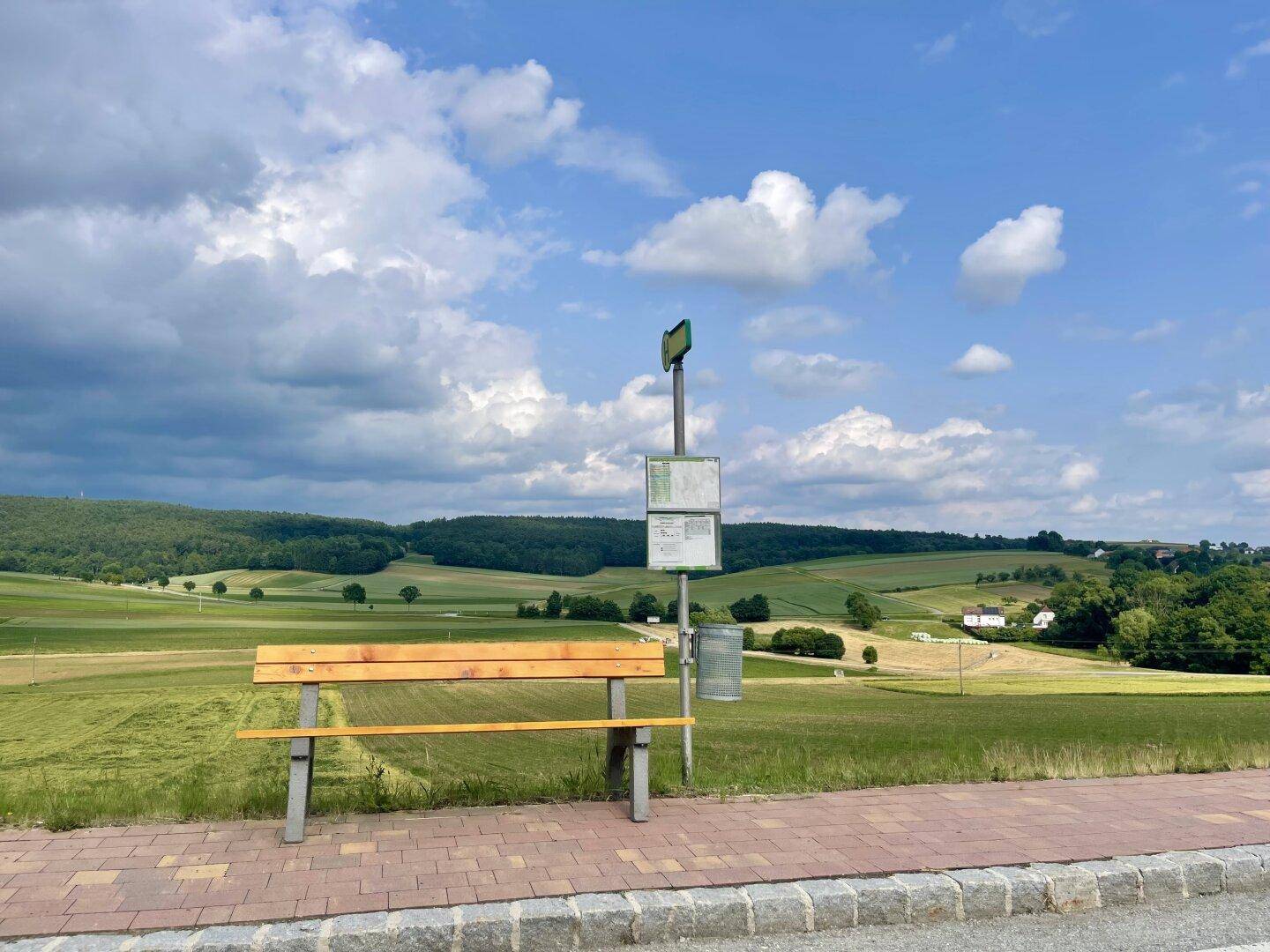 Bushaltestelle mit Sitzbank und weitem Blick über die ländliche, grüne Landschaft.