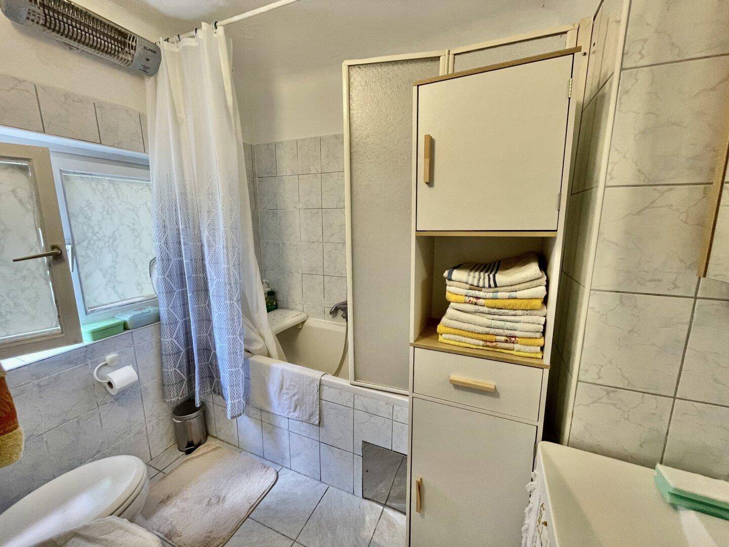 Badezimmer mit Badewanne, Duschvorhang und Fenster, ausgestattet mit hellen Fliesen.