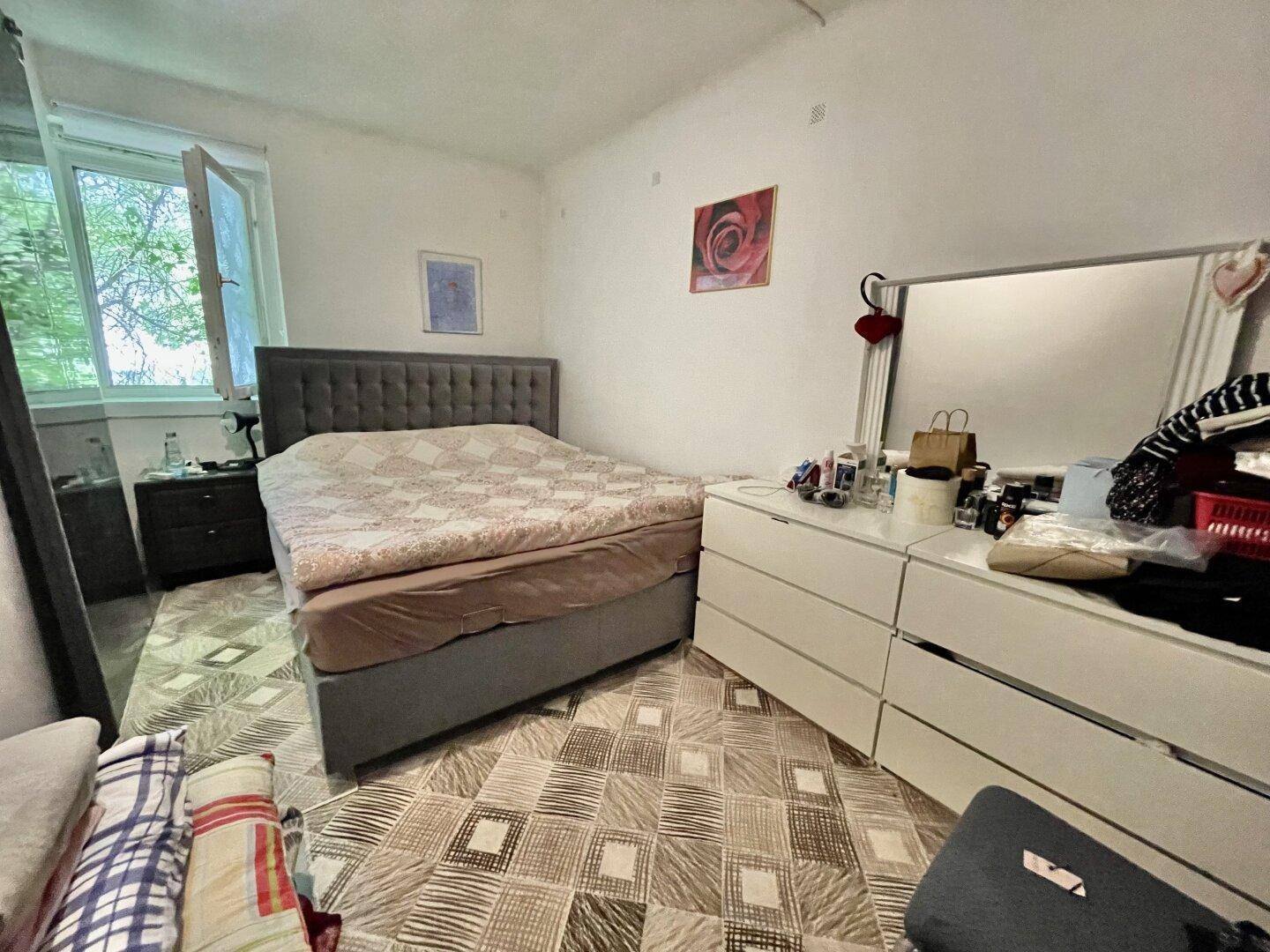 Geräumiges Schlafzimmer mit großem Bett, Kommode und Fenster, das viel Tageslicht hereinlässt.