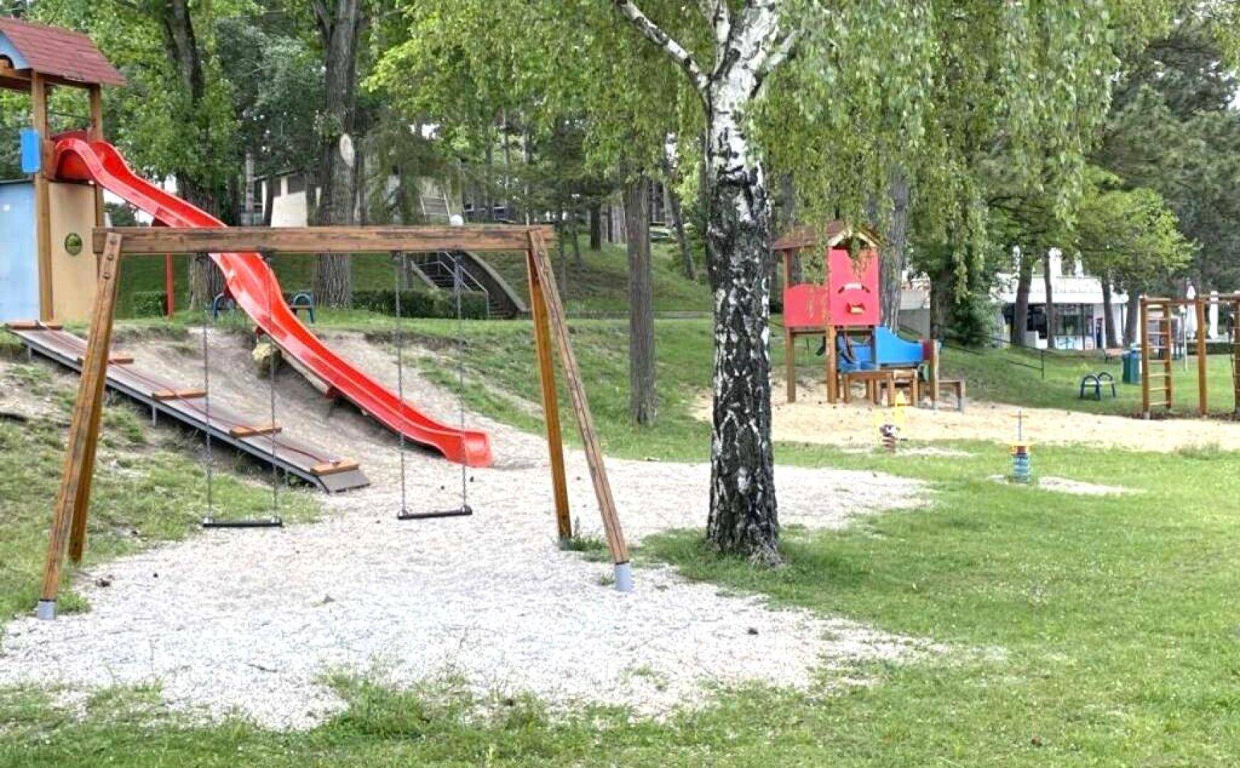 Öffentlicher Spielplatz mit Rutschen, Schaukeln und Sandbereich umgeben von Bäumen.