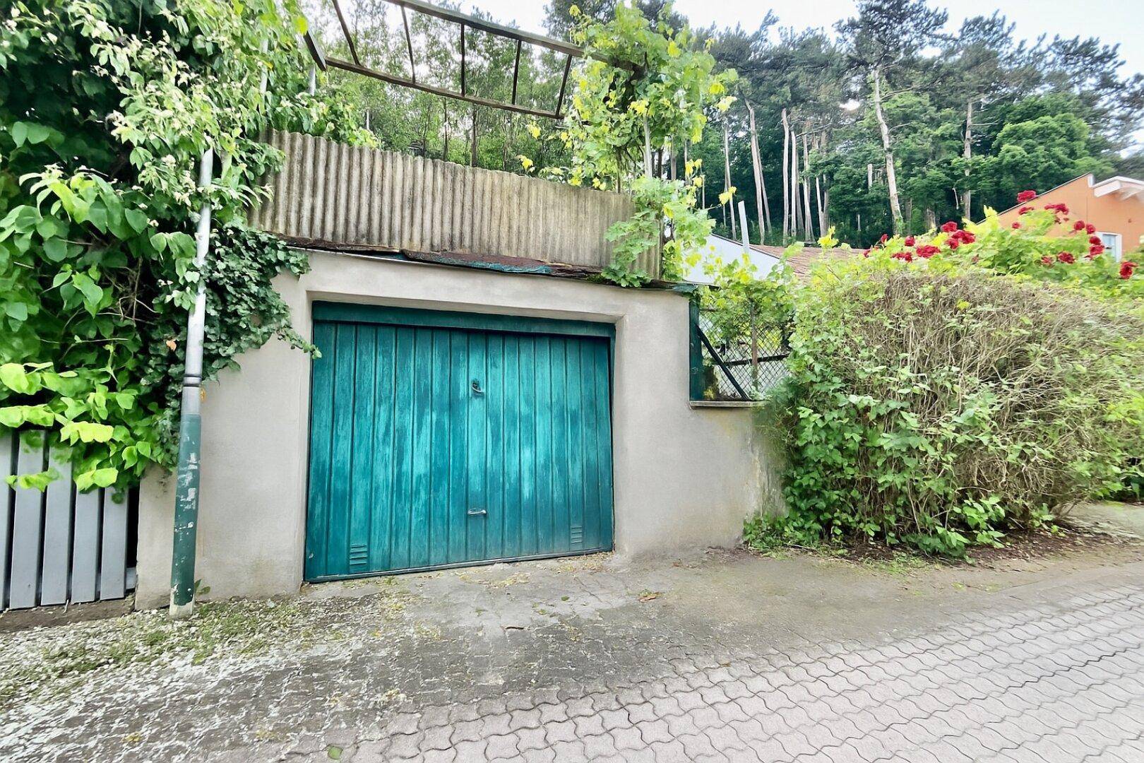 Einzelgarage mit einem grünen Holztor, umgeben von üppigem Grün und Bäumen.