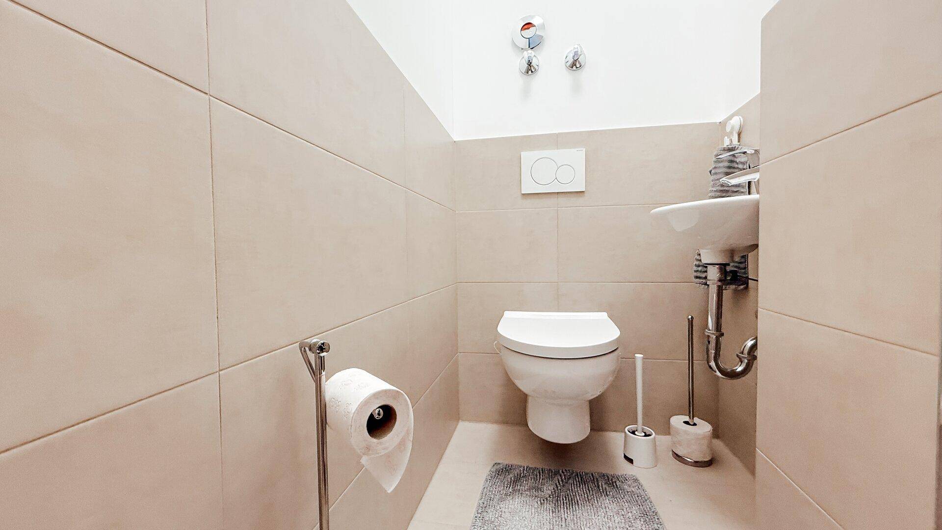 Separates Gäste-WC mit Wand-WC, kleinem Waschbecken und beigefarbenen Fliesen.